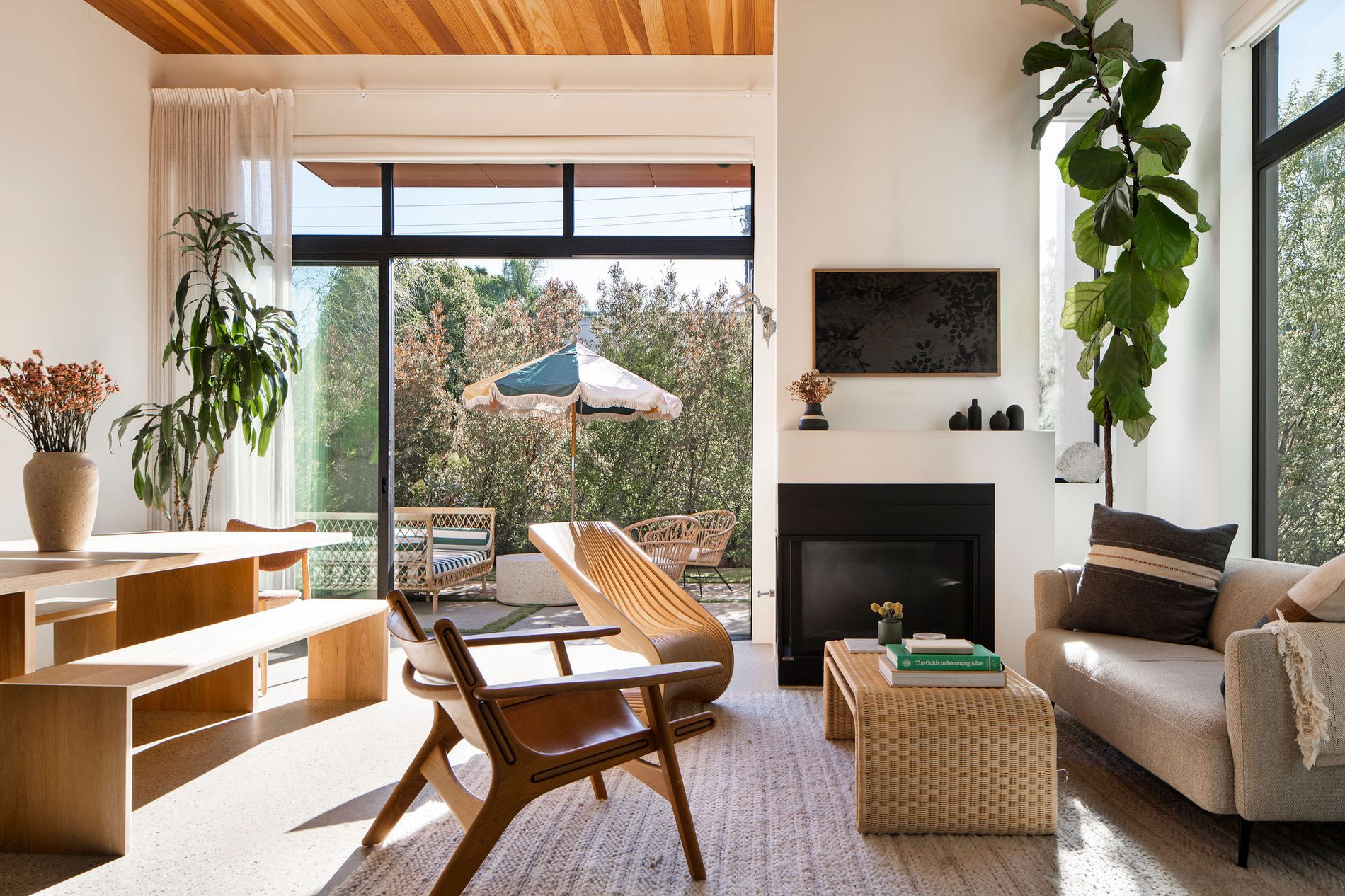 A sunlit living room with a wicker coffee table, leather lounge chair, and fireplace, opening onto a garden patio.